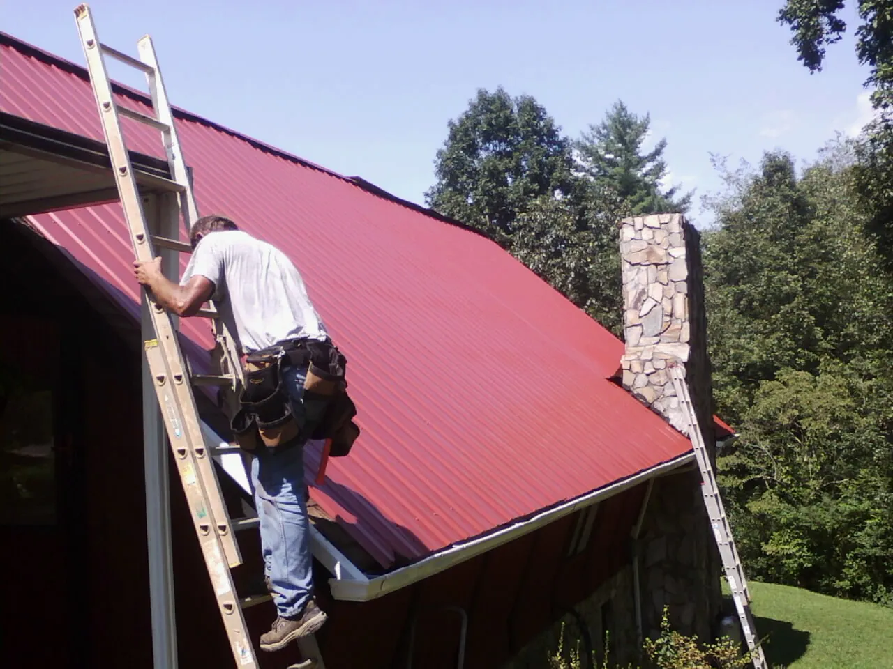 Roofer on a ladder beside a red metal roof and stone chimney — Soffit & Fascia Repair by Roofing Brothers in Waukesha